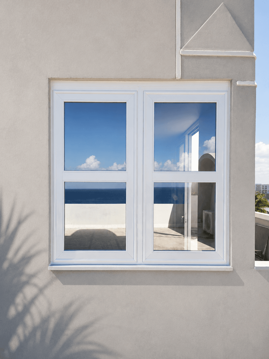 White window in a beige wall reflecting a blue sky and ocean with palm shadows.