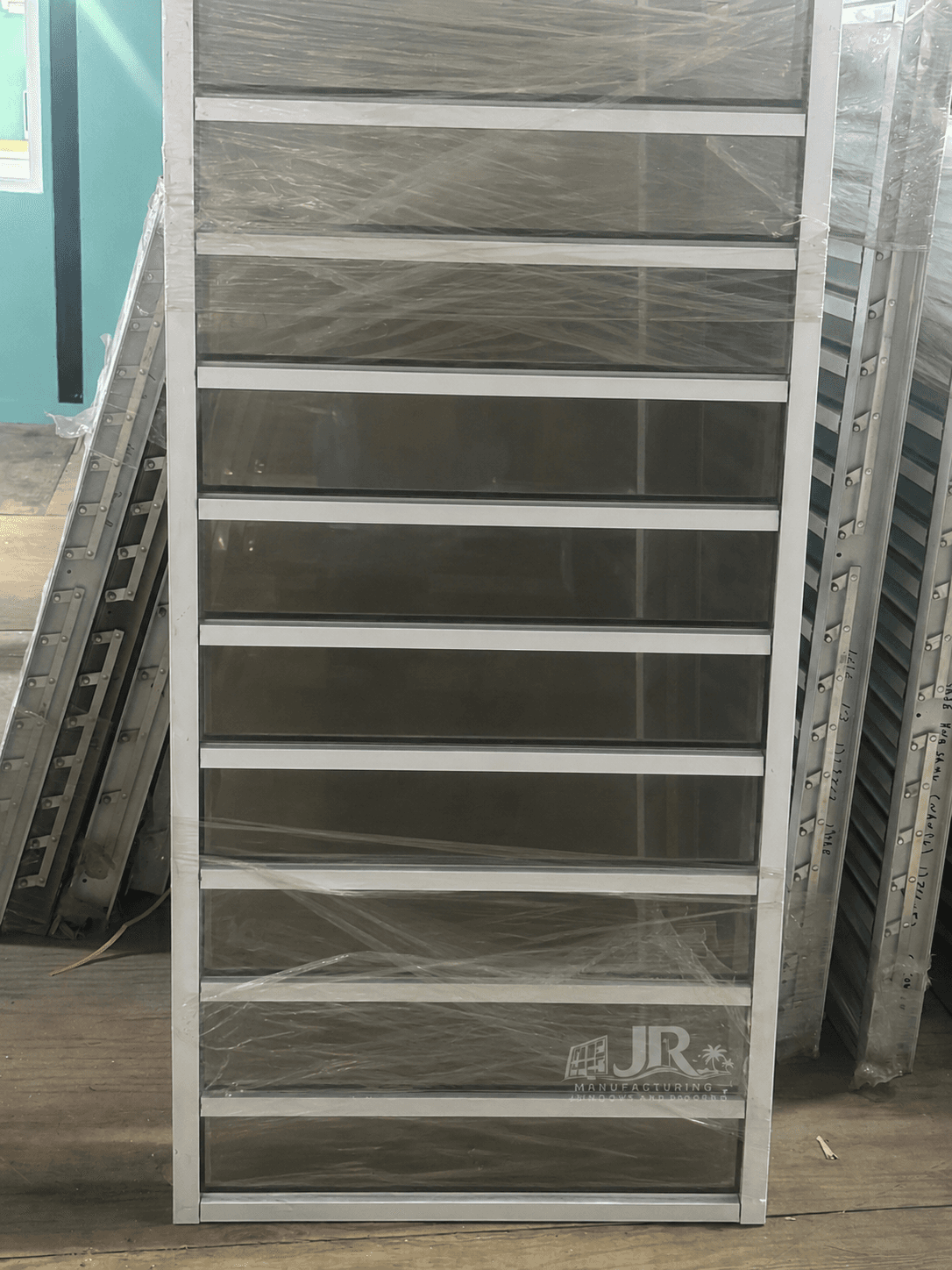 Tall louvered window with white frame and dark glass slats, wrapped in clear protective plastic.