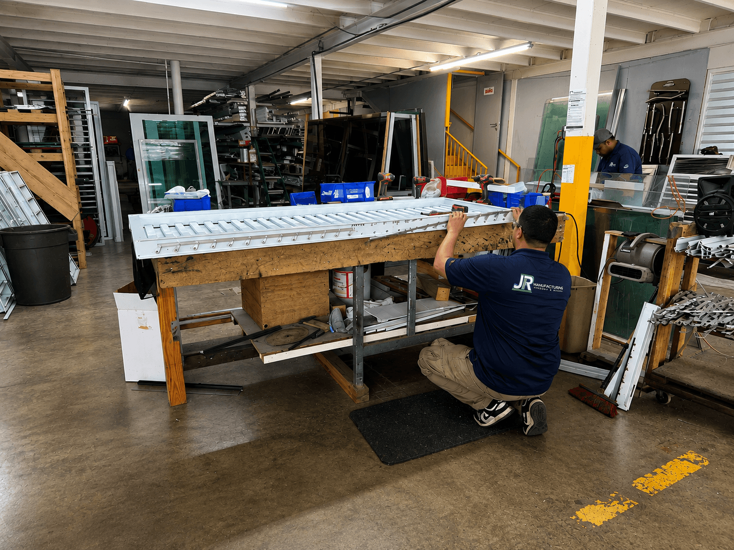 A worker assembles a white shutter on a wooden workbench in a busy manufacturing workshop.