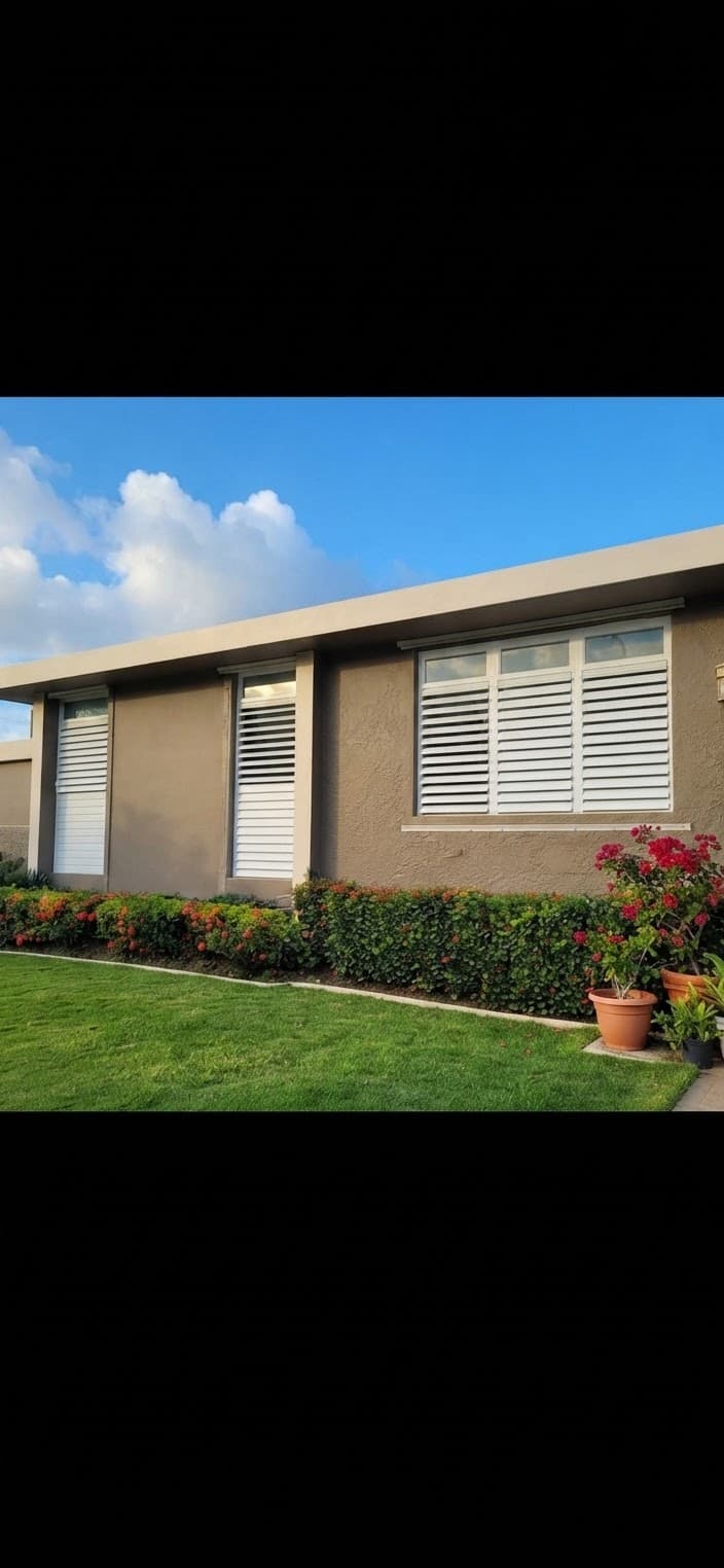 Modern brown house with white shutters, green lawn, and flowering hedges under blue skies.